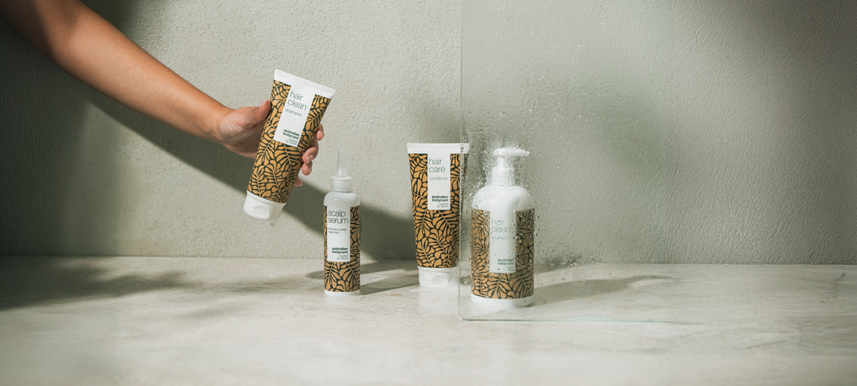 A hand holds a tube of Hair Clean Shampoo from Australian Bodycare in front of a shower screen; Scalp Serum, Hair Care Conditioner and a Hair Clean pump bottle stand below.