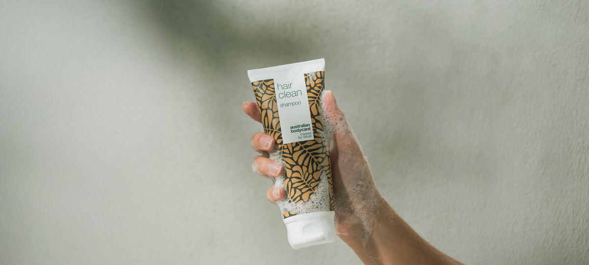 A lathered hand holds a tube of Hair Clean Shampoo from Australian Bodycare in beige packaging in the shower.