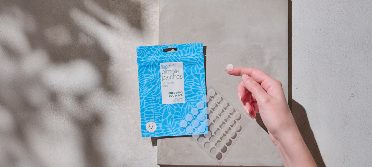 A blue pack of Salicylic Acid Pimple Patches from Australian Bodycare on a stone surface. A hand lifts a single patch off the backing sheet.