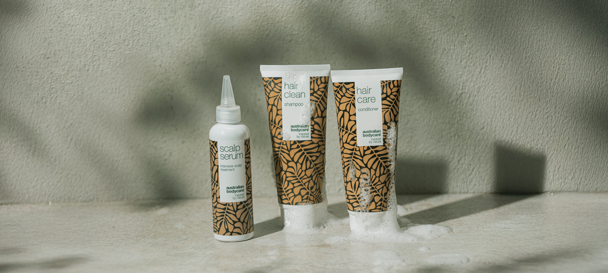 Scalp Serum, Hair Clean Shampoo and Hair Care Conditioner from Australian Bodycare in beige packaging on a stone surface, with foam around the tubes.