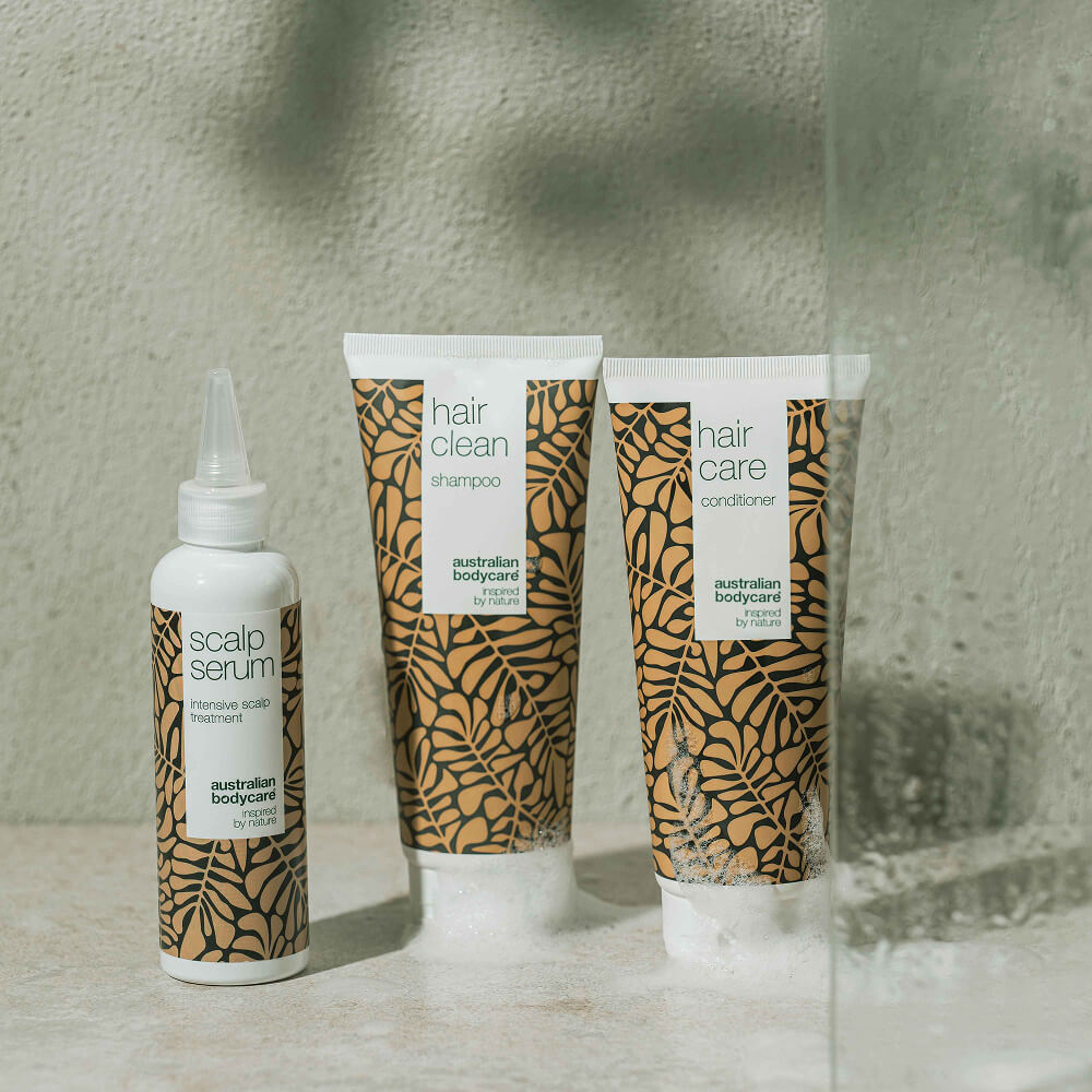 Three Australian Bodycare haircare products – Scalp Serum, Hair Clean Shampoo and Hair Care Conditioner – are standing on a light bathroom surface surrounded by water and foam.